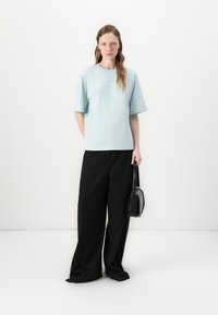 Svetlomodré bavlnené oversized tričko s krátkymi rukávmi, skombinované s čiernymi širokými nohavicami. Modelka drží malú čiernu kabelku.