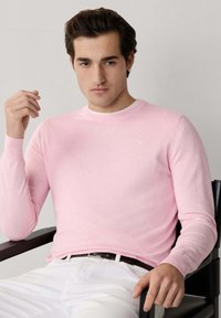 Maglione lavorato a maglia rosa chiaro a maniche lunghe con scollo rotondo, caratterizzato da un logo discreto. Indossato sopra una camicia bianca, abbinato a pantaloni bianchi.