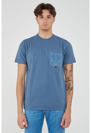 Giovane uomo con braccio destro tatuato che indossa una maglietta blu a girocollo con tasca in denim e jeans azzurri, in piedi davanti a uno sfondo uniforme.