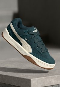 Dark teal sneakers with a suede and mesh upper, white accent stripe, and rubber sole, featuring prominent laces and a cushioned collar.