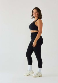 Czarny sportowy biustonosz i legginsy z wysokim stanem, wykonane z elastycznej tkaniny. Modelka nosi białe skarpetki i jasne trampki. Prosty, elegancki design.