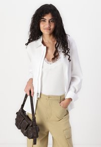 Camicia bianca con zip frontale, sovrapposta a una canotta leggera con dettagli in pizzo. Pantaloni cargo verde oliva. Borsa in pelle marrone.