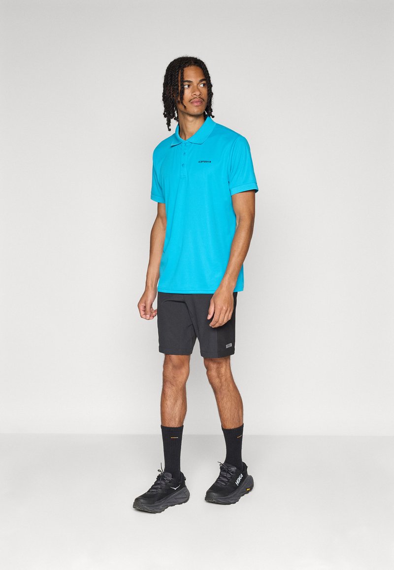 Mann steht vor einem schlichten Hintergrund und trägt ein leuchtend blaues Poloshirt, schwarze Shorts, schwarze Socken und schwarze Sportschuhe.