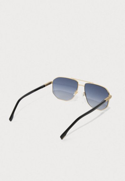 Des lunettes de soleil avec monture dorée, branches noires et verres dégradés bleus, vues de dessus sur un fond blanc.