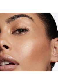 Close-up of a face showing smooth, bronzed skin, defined brows, subtle eye makeup, and glossy lips with a natural shine.