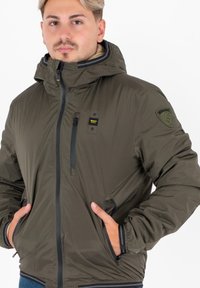 Giacca con cappuccio verde oliva in materiale lucido, dotata di zip frontale, tasche laterali e un logo a toppa testurizzata sulla spalla.