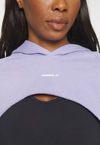 Cropped hoodie viola chiaro con una texture morbida, con cappuccio e logo NEBBIA. Indossato sopra una maglietta nera a collo rotondo.