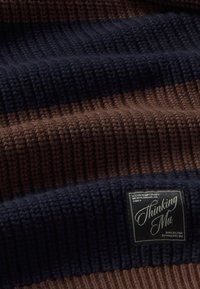 Tissu tricoté rayé en marron et bleu foncé avec une étiquette noire portant l'inscription "Thinking Mu Barcelona".
