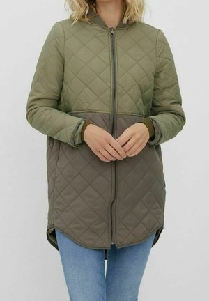 Femme portant une veste matelassée bicolore avec une fermeture éclair à l'avant, debout les mains jointes, portant un jean bleu clair.