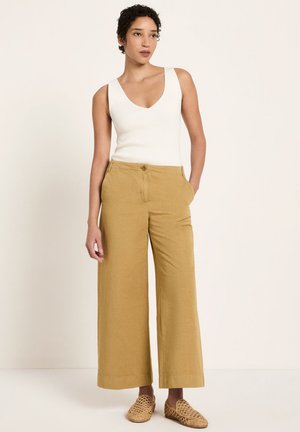 Woman wearing a white sleeveless ribbed top, mustard wide-leg pants, and beige woven slip-on shoes, standing with one hand in pocket.