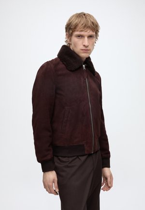 Joven con chaqueta de ante marrón oscuro con cuello de borrego negro y pantalones negros sobre un fondo claro y liso.