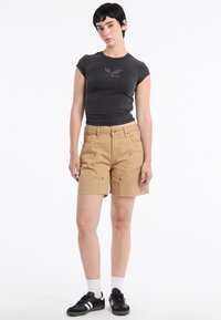 T-shirt nera aderente con grafica di un'aquila, abbinata a pantaloni corti beige fino al ginocchio con molteplici tasche e orlo sfilacciato. Sneakers nere con calzini bianchi.