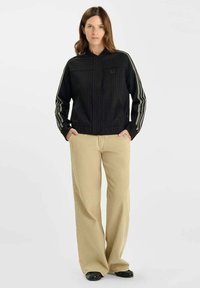 Kvinna som står med händerna i fickorna, klädd i en svart jacka med vita ränder på ärmarna och beige vida byxor.