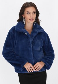 Donna che indossa un morbido giubbotto di pelliccia blu scuro con bottoni decorativi, grandi orecchini e una gonna nera, in posa contro uno sfondo bianco.