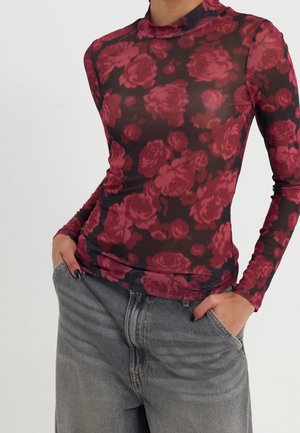 Person iført en tætsiddende, langærmet, gennemsigtig top med røde rosenmønstre over sort, kombineret med løstsiddende grå jeans, hænderne i lommerne.