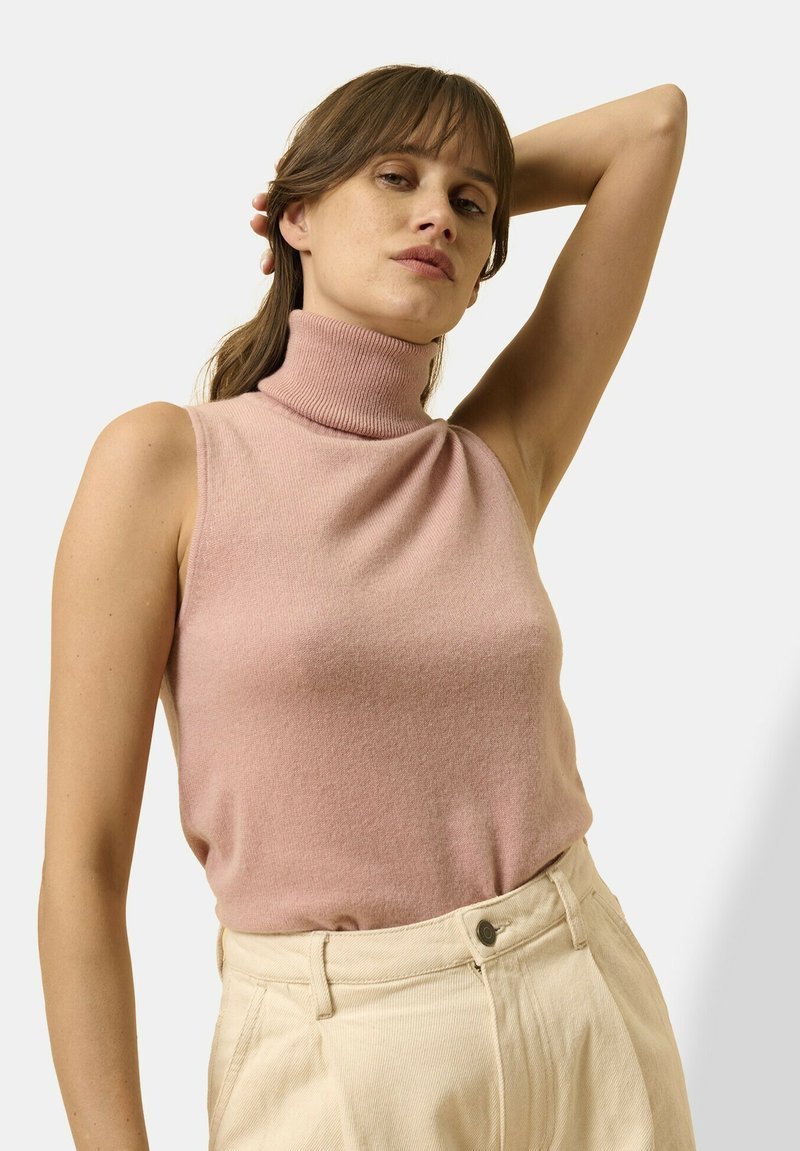 Jasnoróżowy bezrękawnik z golfem wykonany z miękkiego dzianinowego materiału, z dopasowanym krojem i prążkowanym dekoltem. W połączeniu z kremowymi spodniami.