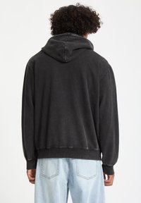 Sudadera negra con un ajuste relajado, que cuenta con una capucha con cordón y puños acanalados. El tejido parece suave y tiene un acabado ligeramente desteñido.