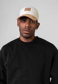 Fehér és bézs sapka puha karimával és bőrpaukkal; fekete crewneck pulóverrel viselve. A semleges színpalettára és a lezser dizájnra helyezve a hangsúlyt.