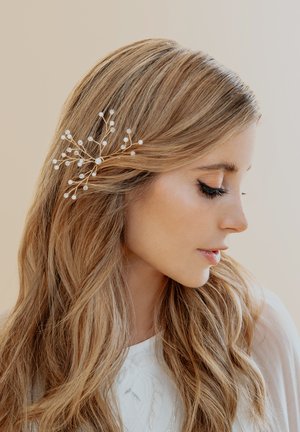 Fermaglio d'oro con accenti di perla, caratterizzato da un design floreale. Indossato su capelli mossi di color castano chiaro, in risalto su uno sfondo neutro.