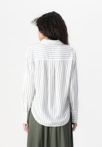 Chemise blanche à rayures verticales noires, à manches longues, avec des poignets à boutons et un pli au dos. Portée par-dessus une jupe unie vert olive.