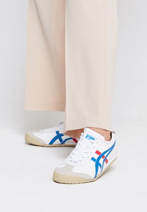 White athletic shoes with blue and red accents, suede toe cap, and textured rubber sole, paired with light beige trousers.