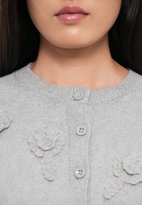 La parte inferiore del viso e il collo di una donna che indossa un maglione grigio chiaro con bottoni, decorato con motivi floreali testurizzati vicino ai bottoni.