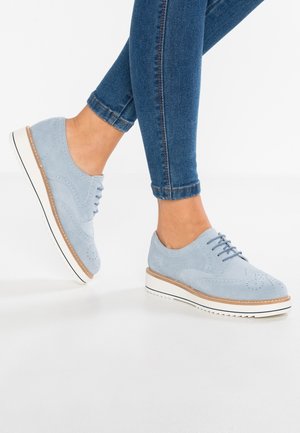 Lysblå ruskinds oxford-sko med brogue-detaljer, hvid gummisål og subtile brune accenter. Båret med mørke denimjeans.