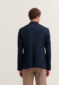 Blazer blu navy con finitura liscia, rever a punta e due spacchi posteriori. Presenta maniche lunghe e polsini con bottoni, abbinato a pantaloni beige.