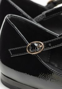 Scarpe in pelle verniciata nera con punta arrotondata, caratterizzate da fasce incrociate e un'applicazione a fibbia ovale dorata. Superficie liscia e lucida con dettagli di cucitura.