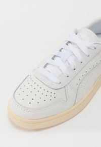 Zapatilla blanca de cuero con diseño perforado, cordones blancos planos y suela de goma crema. Presenta un parche con el logotipo en la lengüeta.
