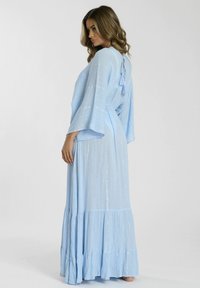 Lång, ljusblå maxiklänning med utsvängda ärmar, knytning i ryggen och subtil glans. Volangkant ger textur och rörelse.
