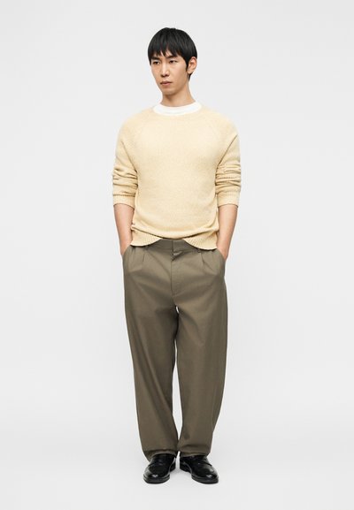 Pull beige clair texturé, sous-vêtement blanc, pantalon ample olive, chaussures noires. Figure isolée les mains dans les poches sur un fond uni.