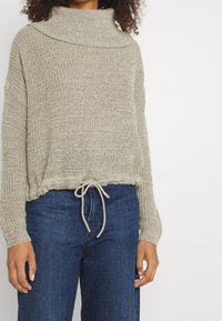 Beige neulepusero, jossa korkea kaulus, kiristysnyöri vyötäröllä ja löysä istuvuus. Yhdistetty tummansinisiin leveälahkeisiin farkkuihin. Texturoitu kangas.