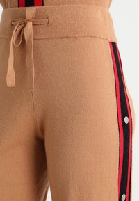 Beżowe dzianinowe spodnie ze sznurkiem w pasie, z bocznym wzorem czerwonych i czarnych pasków oraz srebrnymi zatrzaskami wzdłuż zewnętrznej nogawki.