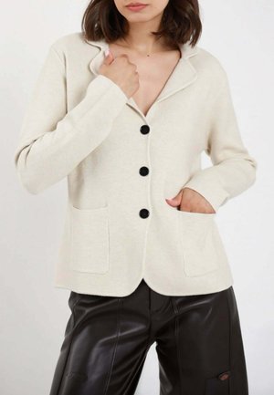 Donna che indossa un cardigan crema con bottoni e due tasche frontali e pantaloni in pelle neri, posando con una mano nella tasca.