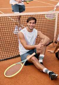 Tennisschläger mit gelbem und grünem Rahmen liegt auf dem orangenen Sandplatz. Ein Mann in weißem ärmellosem Shirt und Shorts sitzt am Netz und lächelt.