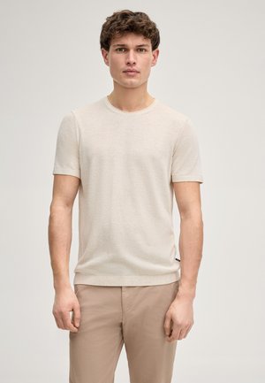 Giovane uomo con capelli corti e ricci castani che indossa una maglietta beige a maniche corte con scollo a girocollo e pantaloni marrone chiaro, in piedi contro uno sfondo semplice.