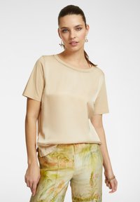 Top beige a maniche corte con scollatura rotonda decorata da cuciture, abbinato a pantaloni chiari a fantasia con motivi tropicali.