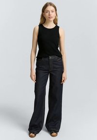 Haut noir sans manches, jean bleu à jambes larges avec poches avant et surpiqûres contrastantes, associé à des sandales marron. Design simple, coupe décontractée.