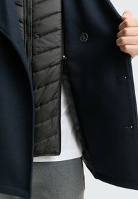 Cappotto esterno in misto lana blu navy con fodera interna trapuntata nera, chiusura con un solo bottone e zip frontale; presenta un design elegante e moderno.