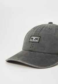 Szürke pamut baseball sapka ívelt karimával és egy fekete hímzett logóval, amely két stilizált hullámot ábrázol. Texturált felület, állítható pánt.