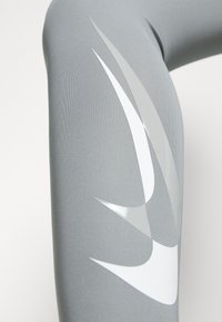 Szare legginsy sportowe z błyszczącymi wzorami w białe i srebrne swoosh. Wykonane z elastycznego materiału o gładkiej teksturze.