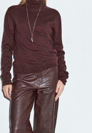 Femme portant un pull à col roulé bordeaux, un pantalon en cuir assorti et un long collier avec un pendentif, debout devant un fond uni.
