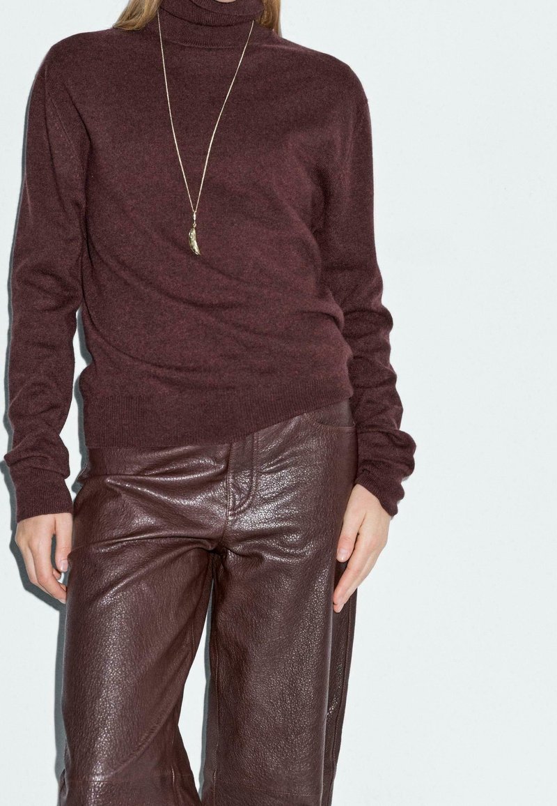 Femme portant un pull à col roulé bordeaux, un pantalon en cuir assorti et un long collier avec un pendentif, debout devant un fond uni.