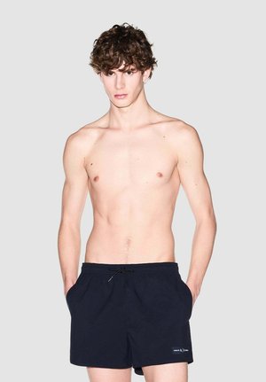 Giovane uomo con capelli ricci che indossa pantaloncini da bagno neri con coulisse, in piedi davanti a uno sfondo chiaro e uniforme.