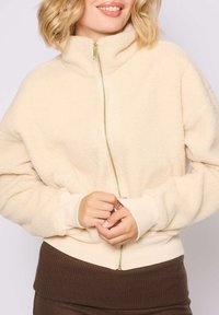 Giacca in pile color crema con colletto alto, zip dorata e polsini a coste. Il tessuto appare morbido e accogliente, in contrasto con l'orlo a coste marrone.
