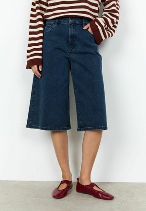 Frau trägt dunkelblaue Jeans-Shorts mit weitem Bein, einen langärmligen Pullover mit bordeauxroten und weißen Streifen sowie bordeauxrote flache Schuhe.