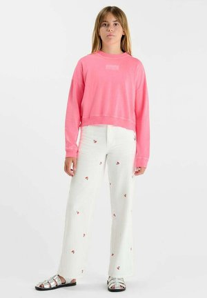 Jonge persoon staand, draagt een roze trui, witte broek met kleine rode patronen en zilveren sandalen met open tenen, tegen een witte achtergrond.