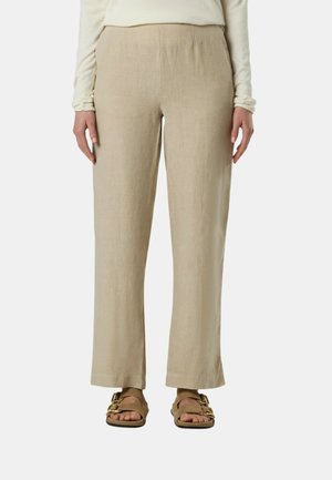 Persona che indossa pantaloni beige a gamba larga, una maglia crema a maniche lunghe e sandali marroni con fibbie dorate, in piedi davanti a uno sfondo semplice.