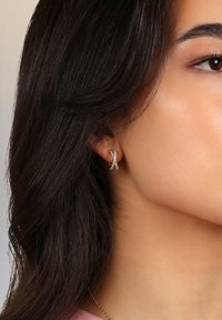 Guldörhänge med vridet ribbdesign, dekorerad med små klara ädelstenar, visad på en models öra mot en neutral bakgrund.